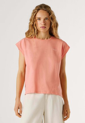 Donna con capelli biondi ondulati che indossa una maglietta a maniche corte color corallo e pantaloni beige chiaro, in piedi davanti a uno sfondo semplice.