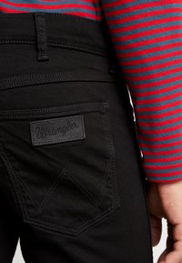 Melnas denima bikses ar izteiksmīgu Wrangler logo uz aizmugurējā kabatas, detalizētas ar kontrastējošu šuvju un šauras griezuma dizainu.