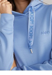 Felpa con cappuccio azzurro chiaro indossata da una persona, caratterizzata da un cordoncino marchiato con il testo "JOOP!" e un logo "JOOP!" ricamato sul petto.