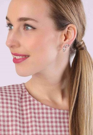 Silberne Ohrringe mit Herzdesign, verziert mit einem kleinen Edelsteinakzent. Das Modell trägt eine pink karierte Bluse mit Rundhalsausschnitt.