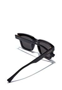 Mustad musta raami ja tumedate läätsedega prillid, kaldes küljele, näidates kuldset "HAWKERS" brändi nime prillide templitel.