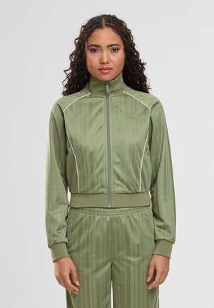 Frau mit lockigem Haar, die eine olivgrüne Trainingsjacke mit Reißverschluss und passende Hose trägt, steht vor einem einfarbigen hellgrauen Hintergrund.