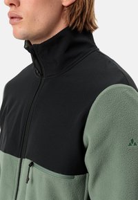 Giacca in pile con la parte superiore nera e la parte inferiore verde. Presenta un colletto alto, una tasca sul petto con zip e un tessuto strutturato.