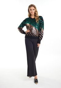 Pulover negru cu dungi diagonale din paiete verzi și bronz, asortat cu pantaloni negri largi și pantofi negri cu accente metalice.