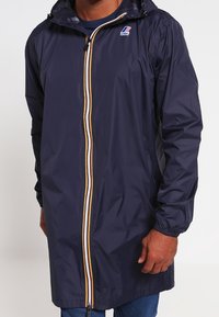 Laivastonsininen vedenpitävä takki, jossa on vetoketjullinen etuosa, kelta-valkoiset raidalliset yksityiskohdat, joustavat hihansuut ja huppu, valmistettu kevyestä, kiiltävästä kankaasta.