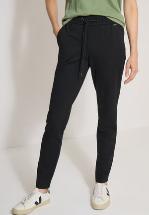 Personne portant un pantalon noir à cordon, un t-shirt vert et des baskets blanches, debout les mains dans les poches.