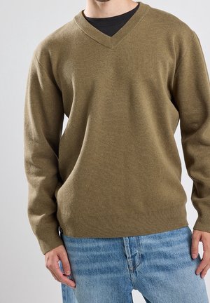 Uomo che indossa un maglione verde oliva a scollo a V sopra una camicia nera e jeans azzurri sullo sfondo semplice.