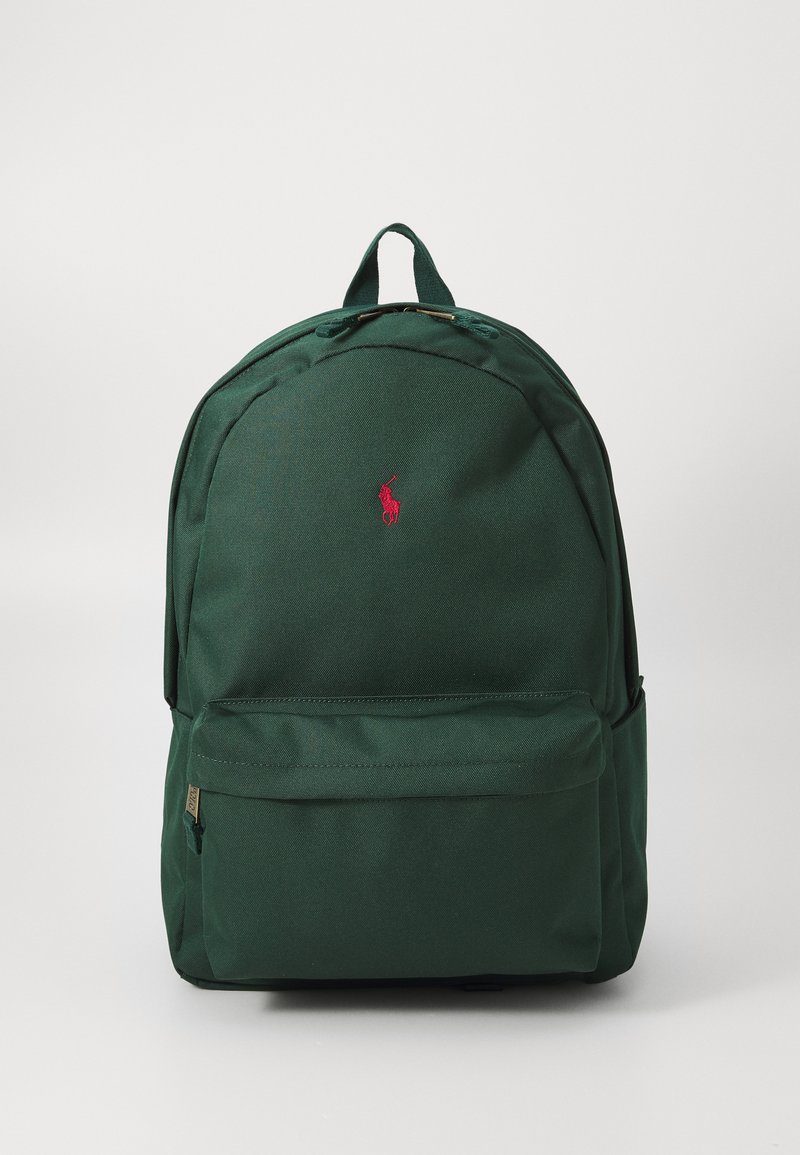 Mochila verde hecha de un tejido texturizado, con una parte superior redondeada, bolsillo frontal y un pequeño logo rojo. Tiene correas ajustables y un asa para llevarla.