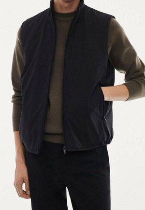 Personne portant un gilet sans manches noir sur un pull olive et un pantalon noir, une main dans la poche du gilet, debout devant un fond uni.