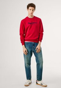 Rød sweatshirt med marineblå logo, parret med adskillige slidte blå jeans. Casual sneakers har en blanding af hvid, creme og sorte accenter.