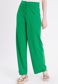 Femme portant un pantalon large taille haute vert avec une taille boutonnière, associé à des sandales à plateforme dorées sur un fond blanc.