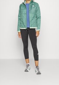 Zelená prošívaná bunda se světlým lemem, modrý top, černé legíny a vícebarevné boty, s zipovým zapínáním a stojatým límcem.