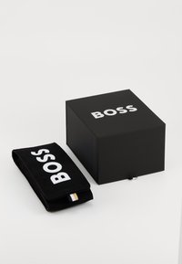 Caixa preta com o logótipo branco "BOSS" ao lado de um saco preto com o mesmo logótipo, com uma pequena etiqueta de destaque em castanho e branco.