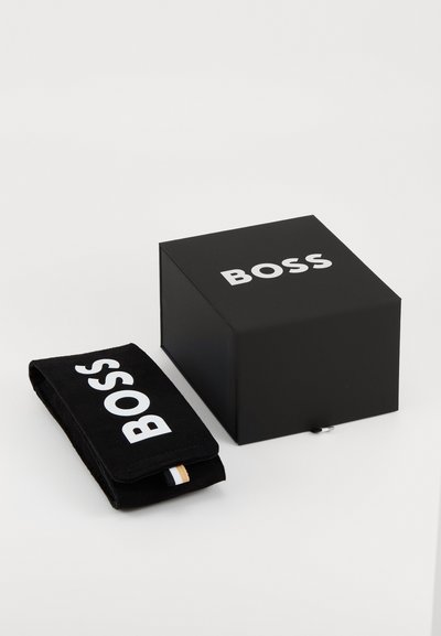 Boîte noire avec le logo "BOSS" en blanc. À côté, une pochette noire en matériau doux, également avec le logo "BOSS", ornée d'une étiquette accent colorée.