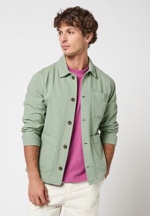 Joven con chaqueta abotonada verde salvia sobre camisa morada y pantalones blanco hueso, mirando hacia la izquierda sobre un fondo liso.