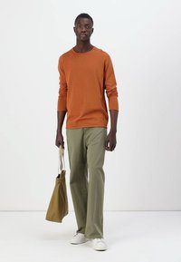 Oranges Langarmhemd, grüne Hose, weiße Sneaker, hält eine beige Tragetasche. Einfaches Design, normale Passform, Baumwollstoff, neutraler Hintergrund.