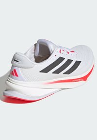 Chaussure de sport blanche avec des rayures noires, des détails gris et une semelle rouge. Présente un dessus en maille texturée et une semelle intermédiaire rembourrée.