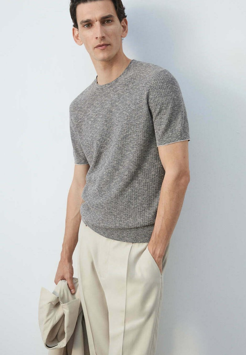 Homme portant un pull en maille gris à manches courtes et un pantalon beige, tenant une veste beige dans la main gauche sur un fond uni.