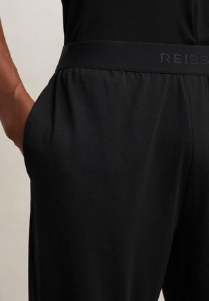 Persona che indossa pantaloni neri con vita elastica etichettata "REISS", mano nella tasca laterale, mostrata dalla vita a metà coscia.