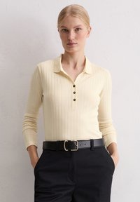 Blonde Frau mit einem cremefarbenen, gerippten Langarmoberteil mit schwarzen Knöpfen, schwarzen Hosen und einem schwarzen Gürtel mit einer goldenen Schnalle. Hände in den Taschen.