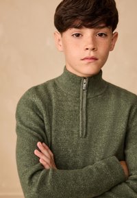 Grøn, lynlåstrøje med høj krave og ribbet kant. Materialet virker blødt med en tekstureret overflade. Modellen har armene krydsede.