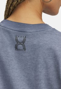 Sudadera de algodón azul con un logotipo gráfico negro en la parte posterior, que muestra "UNDER ARMOUR" y líneas estilizadas, con un cuello acanalado.