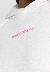 Jasnoszary hoodie z wielokolorowymi plamkami. Na froncie znajduje się różowe logo "new balance" wyszyte na stałe. Miękki materiał oraz luźny krój.