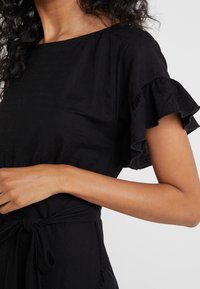 Mujer vestida con un vestido negro de mangas cortas con volantes y un cinturón atado en la cintura, mostrando el torso superior y parte del brazo izquierdo contra un fondo sencillo.