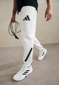 Bílé sportovní kalhoty s černými pruhy a logem adidas, doplněné o bílé tenisky s černými akcenty. Osoba drží skákací provaz.