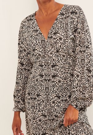 Femme portant une robe à manches longues et col en V avec des motifs floraux et abstraits noirs et beiges, manches rassemblées aux poignets.