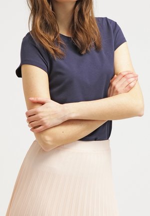 Vrouw draagt een marineblauw T-shirt met korte mouwen en een lichtroze geplooide rok, armen gekruist, tegen een effen lichte achtergrond.