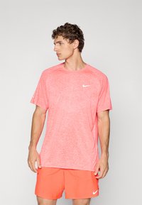 Hombre vestido con una camiseta Nike en color coral jaspeado y pantalones cortos Nike en color coral sólido, de pie contra un fondo gris claro liso, mirando hacia la izquierda.