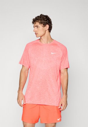 Hombre vestido con una camiseta Nike en color coral jaspeado y pantalones cortos Nike en color coral sólido, de pie contra un fondo gris claro liso, mirando hacia la izquierda.