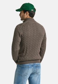 Brauner Kabelstrickpullover mit hohem Kragen, gerippten Bündchen und Saum, kombiniert mit hellblauen Jeans. Dunkelgrüne Mütze mit einem Etikett und Logodetail.