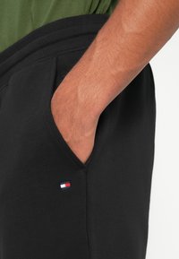 Sorte joggebukser med en glatt tekstur, elastisk midje og sidelommer. Inkluderer en liten brodert logo i rødt, hvitt og blått.