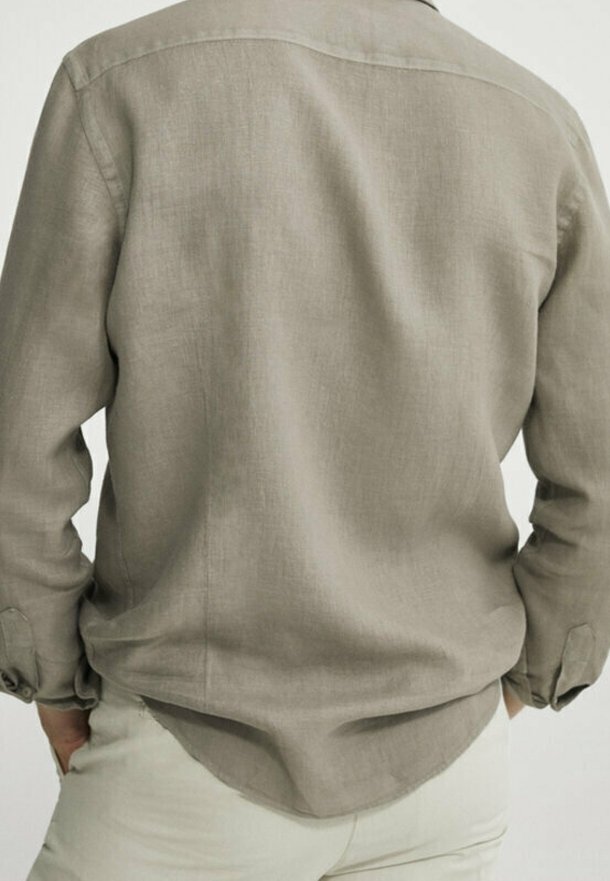Homme portant une chemise beige ample à manches longues portée sortie sur un pantalon clair, vu de dos avec les mains dans les poches.