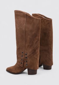 Botas de ante marrón hasta la rodilla con punta afilada, que cuentan con una superficie texturizada y detalles de ojales negros en el lado. Tacón de madera apilada.