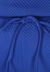 Blauer Strickstoff mit einem strukturierten Fischgrätenmuster. Ein Tunnelzug ist an der Taille gebunden und fügt dem Design funktionale Details hinzu.