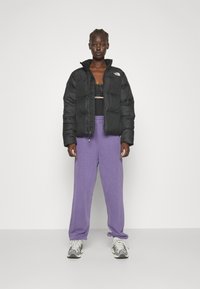 Giacca a vento nera con zip frontale e colletto alto, indossata sopra un top nero, abbinata a pantaloni sportivi viola larghi e scarpe grigie.