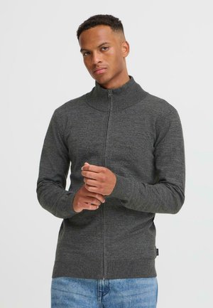 Mann i en grå zip-up genser står mot en enkel bakgrunn, ser selvsikkert på kameraet med hendene sammenfoldet.