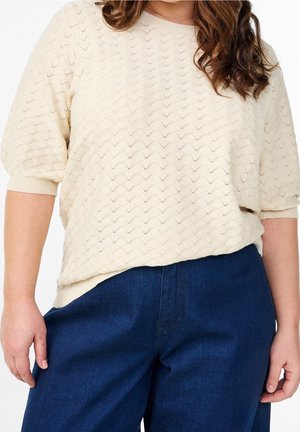 Femme portant un pull beige texturé à manches courtes et un pantalon taille haute bleu foncé, avec des cheveux bruns ondulés visibles.