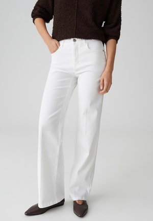 Persoon draagt een witte wijde broek met hoge taille, een zwart textuur langemouwen shirt en donkere puntige schoenen, staat met één hand in de zak.