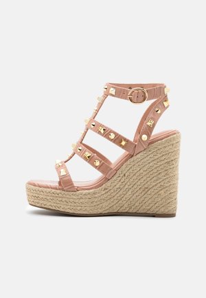 Sandalia cuña en faux cuero rosa claro con tachuelas doradas, con múltiples correas y una plataforma estilo espadrille trenzada.