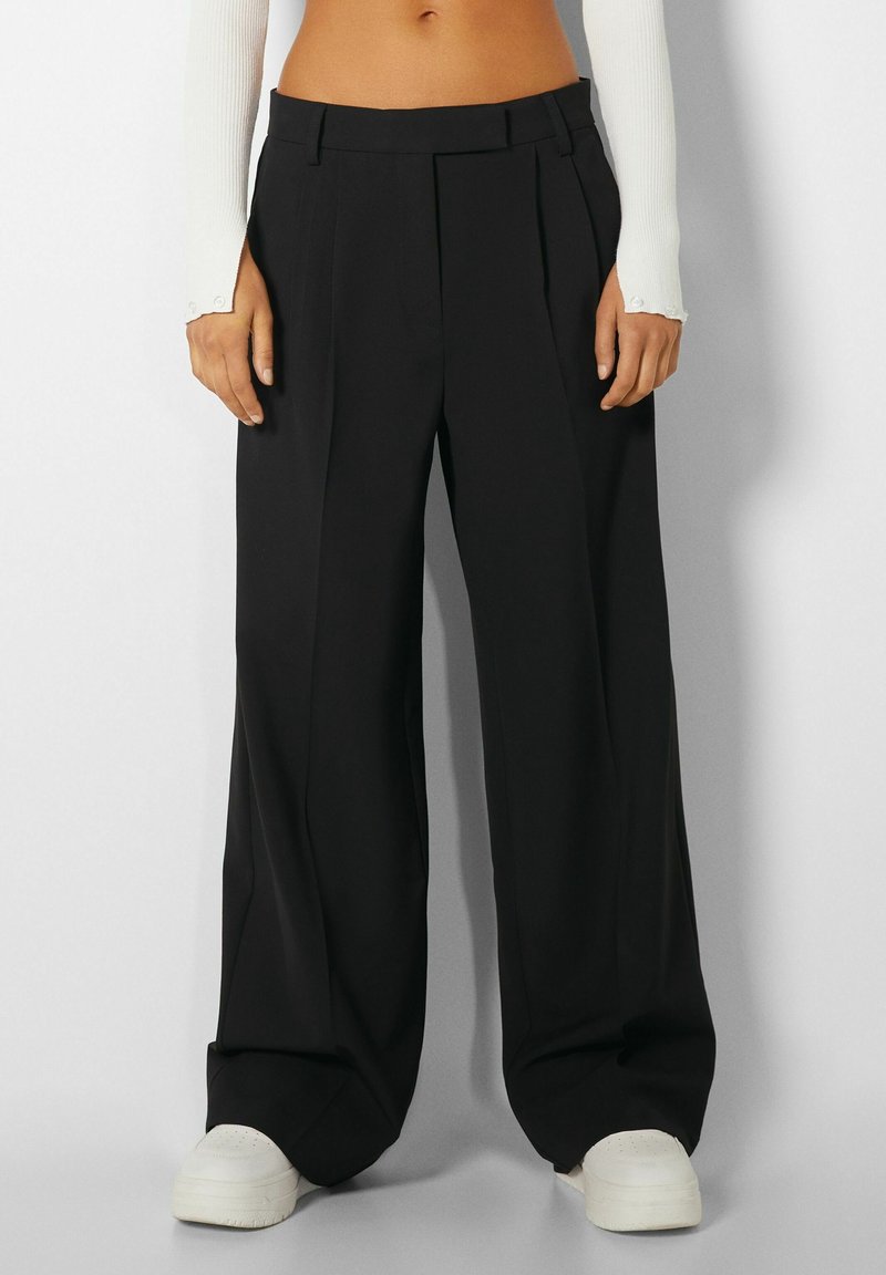 Schwarze weite Hosen mit hoher Taille, plissiertem Design und glattem Stoff. Abgeschlossen mit einem Gürtelschlauf und kombiniert mit weißen Sneakers.