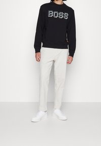 Sudadera negra con el logo "BOSS" en un diseño pixelado, combinada con pantalones beige claros y zapatillas blancas, todo sobre un fondo liso.