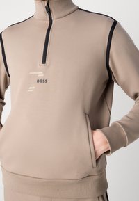 Muž nosí béžovou mikinu na zip s černým lemováním a logem BOSS na hrudi, ruka v přední kapse.