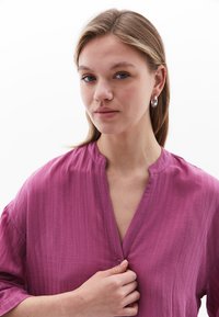 Blusa fucsia con bottoni e finitura testurizzata, caratterizzata da un colletto senza collo e maniche larghe e ampie. La modella tiene un bottone al collo.