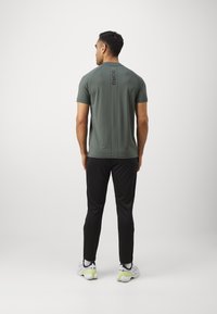 Szürke sport póló rövid ujjakkal, fekete függőleges logóval a hátsó részén, fekete nadrággal és színes sportcipővel párosítva.