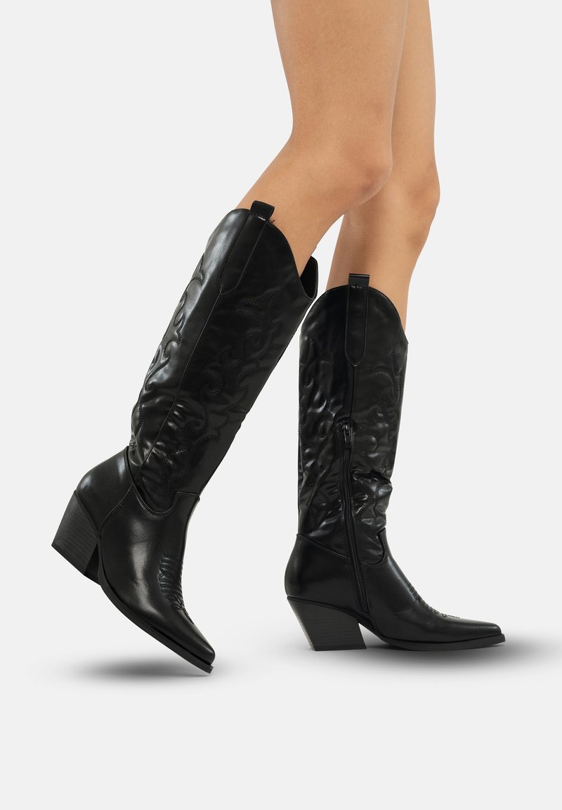Botas de vaquero de cuero sintético negro con punta puntiaguda, tacón medio, costuras decorativas y cremalleras laterales. Textura suave con patrón acentuado.