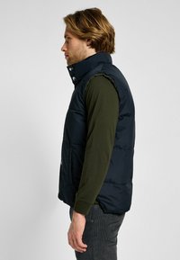 Gilet imbottito blu navy, caratterizzato da un alto colletto, dettagli con bottoni sul davanti e tasche laterali. Indossato sopra una camicia verde a maniche lunghe, con una texture liscia.